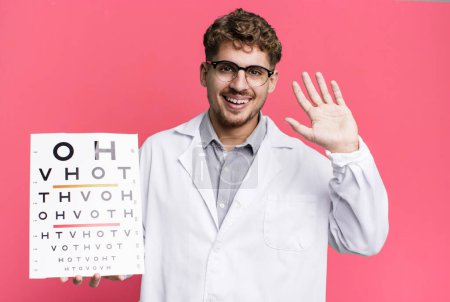 Foto de Young adult caucasian man smiling happily, waving hand, welcoming and greeting you. optical vision test concept - Imagen libre de derechos