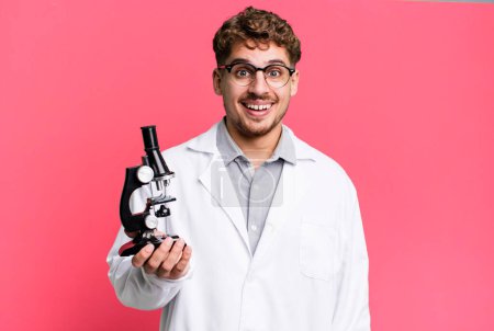Téléchargez les photos : Young adult caucasian man looking happy and pleasantly surprised. scients laboratory student with a microscope concept - en image libre de droit