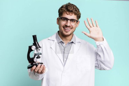 Téléchargez les photos : Young adult caucasian man smiling happily, waving hand, welcoming and greeting you. scients laboratory student with a microscope concept - en image libre de droit