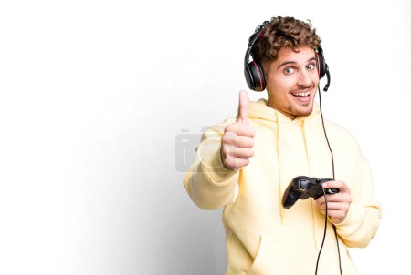 Foto de Young adult caucasian man feeling proud,smiling positively with thumbs up with headset and a controller. gamer concept - Imagen libre de derechos
