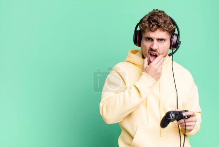 Foto de Young adult caucasian man with mouth and eyes wide open and hand on chin with headset and a controller. gamer concept - Imagen libre de derechos