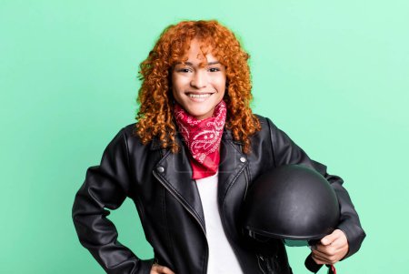 Téléchargez les photos : Cheveux roux jolie femme souriant joyeusement avec une main sur la hanche et confiant. concept de motocycliste - en image libre de droit