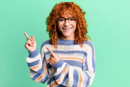 Photo for Redhair pretty woman smiling happily and pointing to side and upwards with both hands showing object in copy space - Royalty Free Image