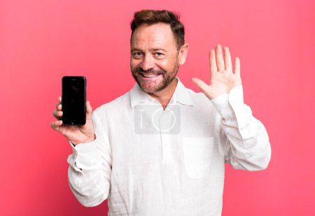Téléchargez les photos : Middle age man smiling happily, waving hand, welcoming and greeting you. showing a smartphone - en image libre de droit