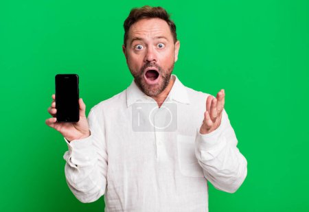 Foto de Middle age man amazed, shocked and astonished with an unbelievable surprise. showing a smartphone - Imagen libre de derechos