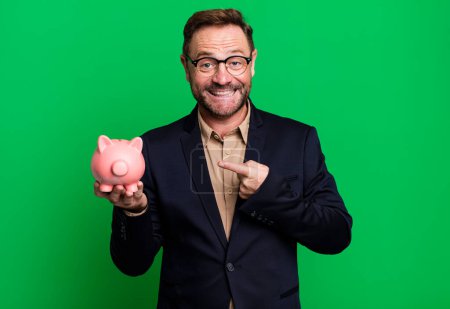 Téléchargez les photos : Middle age man looking excited and surprised pointing to the side with a piggy bank. business concept - en image libre de droit