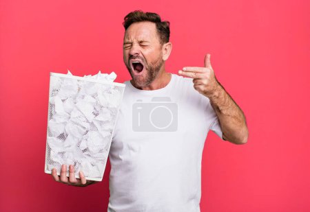 Foto de Middle age man looking unhappy and stressed, suicide gesture making gun sign. paper balls trash basket - Imagen libre de derechos