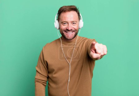 Téléchargez les photos : Middle age man pointing at camera choosing you. listening music with a headphones - en image libre de droit