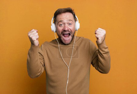 Téléchargez les photos : Middle age man feeling shocked,laughing and celebrating success. listening music with a headphones - en image libre de droit