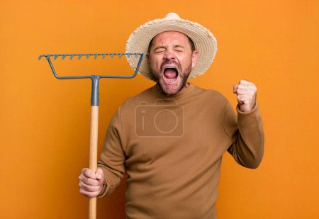 Foto de Middle age man shouting aggressively with an angry expression. farmer with a rake - Imagen libre de derechos