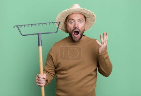 Téléchargez les photos : Middle age man feeling extremely shocked and surprised. farmer with a rake - en image libre de droit
