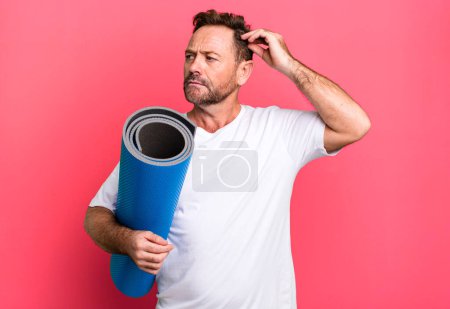 Foto de Middle age man smiling happily and daydreaming or doubting. with a yoga matt. fitness concept - Imagen libre de derechos