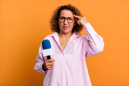 Foto de Una mujer de mediana edad sintiéndose confundida y perpleja, mostrando que estás loca. presentador de televisión con un concepto de micrófono - Imagen libre de derechos