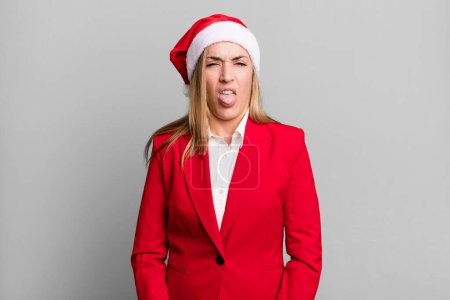 Foto de Bonita mujer rubia sintiéndose disgustada e irritada y la lengua hacia fuera. concepto de Navidad y sombrero de santa - Imagen libre de derechos