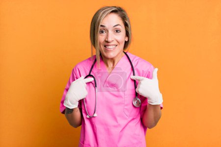 Photo for Pretty blonde woman feeling happy and pointing to self with an excited. nurse concept - Royalty Free Image
