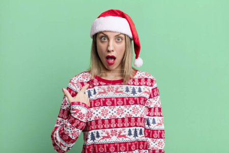 Téléchargez les photos : Pretty blonde woman looking shocked and surprised with mouth wide open, pointing to self. christmas and santa hat concept - en image libre de droit
