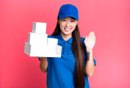 Téléchargez les photos : Pretty asian woman smiling happily, waving hand, welcoming and greeting you. delivery packages concept - en image libre de droit