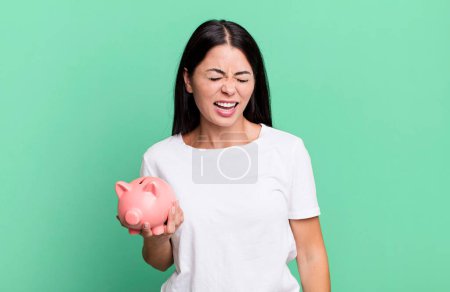Photo for Hispanic pretty woman shouting aggressively, looking very angry with a piggy bank - Royalty Free Image