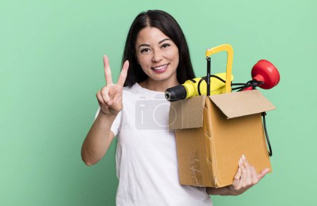 Photo for Hispanic pretty woman smiling and looking happy, gesturing victory or peace with a tool box - Royalty Free Image