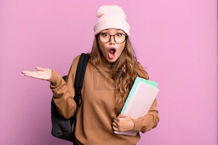 Photo for Hispanic pretty woman looking surprised and shocked, with jaw dropped holding an object. university student concept - Royalty Free Image