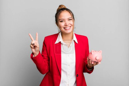 Foto de Hispanic pretty woman smiling and looking friendly, showing number two with a piggy bank - Imagen libre de derechos