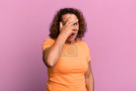 Foto de Mujer hispana de mediana edad que parece sorprendida, asustada o aterrorizada, cubriendo la cara con la mano y mirando entre los dedos - Imagen libre de derechos