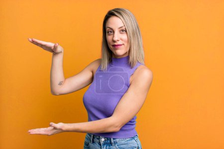 Photo for Blonde adult woman smiling, feeling happy, positive and satisfied, holding or showing object or concept on copy space - Royalty Free Image