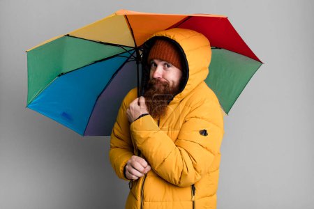Téléchargez les photos : Longue barbe et cheveux roux homme cool avec parapluie, chapeau et un manteau. concept de motte et d'hiver - en image libre de droit