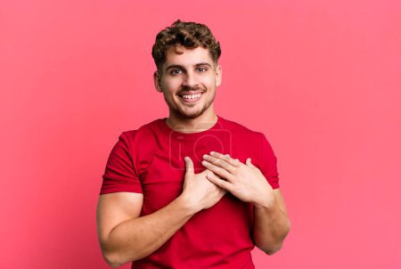 Téléchargez les photos : Young adult caucasian man feeling romantic, happy and in love, smiling cheerfully and holding hands close to heart - en image libre de droit