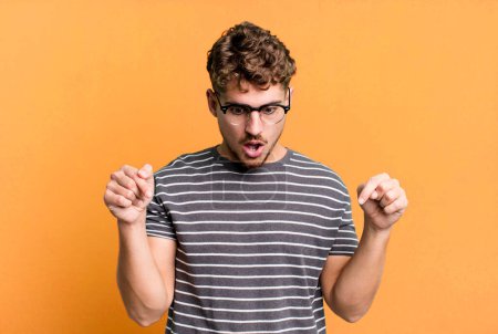 Photo for Young adult caucasian man feeling shocked, open-mouthed and amazed, looking and pointing downwards in disbelief and surprise - Royalty Free Image