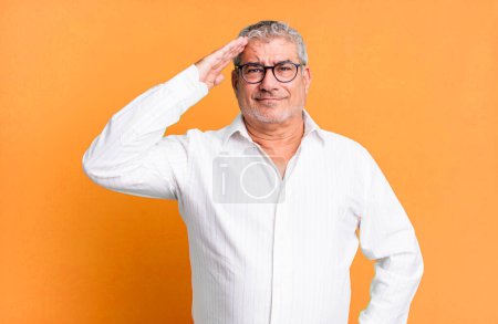 Téléchargez les photos : Homme âgé d'âge moyen saluant la caméra avec un salut militaire dans un acte d'honneur et de patriotisme, faisant preuve de respect - en image libre de droit