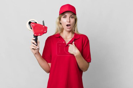Foto de Caucasian blonde woman looking shocked and surprised with mouth wide open, pointing to self. shipping packer concept - Imagen libre de derechos