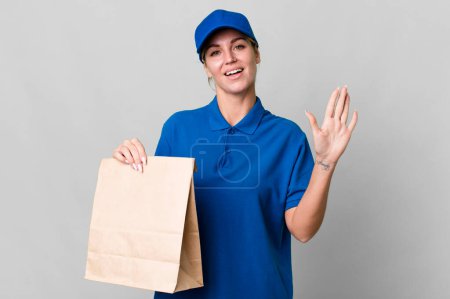 Foto de Caucasian blonde woman smiling happily, waving hand, welcoming and greeting you. paper bag delivery concept - Imagen libre de derechos