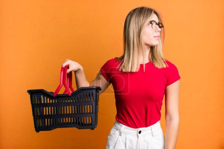 Photo for Young pretty woman on profile view thinking, imagining or daydreaming. empty shopping basket concept - Royalty Free Image