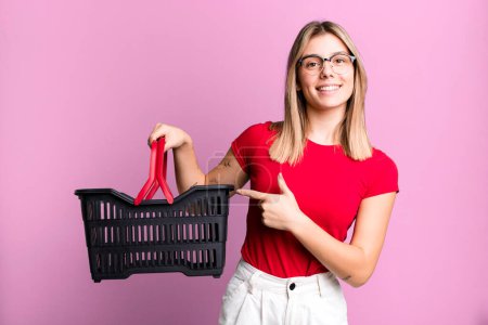 Photo for Young pretty woman looking excited and surprised pointing to the side. empty shopping basket concept - Royalty Free Image