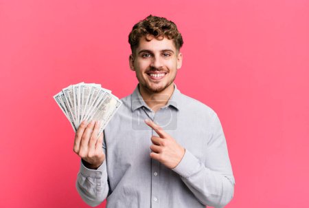 Téléchargez les photos : Young adult caucasian man smiling cheerfully, feeling happy and pointing to the side. dollar bank notes concept - en image libre de droit