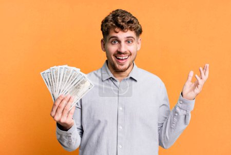 Foto de Young adult caucasian man feeling happy, surprised realizing a solution or idea. dollar bank notes concept - Imagen libre de derechos