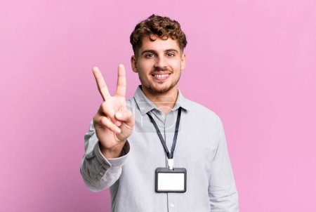 Foto de Young adult caucasian man smiling and looking happy, gesturing victory or peace. blank accreditation pass card id concept - Imagen libre de derechos