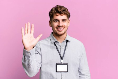 Foto de Young adult caucasian man smiling happily, waving hand, welcoming and greeting you. blank accreditation pass card id concept - Imagen libre de derechos