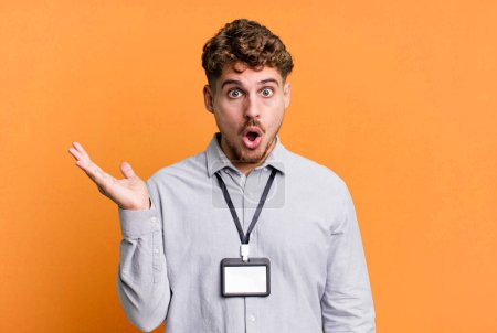 Foto de Young adult caucasian man looking surprised and shocked, with jaw dropped holding an object. blank accreditation pass card id concept - Imagen libre de derechos