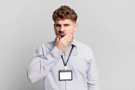 Foto de Young adult caucasian man with mouth and eyes wide open and hand on chin. blank accreditation pass card id concept - Imagen libre de derechos