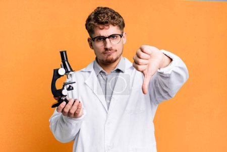 Foto de Young adult caucasian man feeling cross,showing thumbs down. scients laboratory student with a microscope concept - Imagen libre de derechos