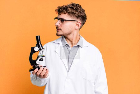 Photo for Young adult caucasian man on profile view thinking, imagining or daydreaming. scients laboratory student with a microscope concept - Royalty Free Image