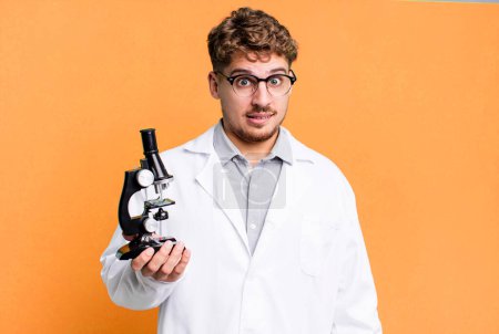 Foto de Young adult caucasian man looking puzzled and confused. scients laboratory student with a microscope concept - Imagen libre de derechos