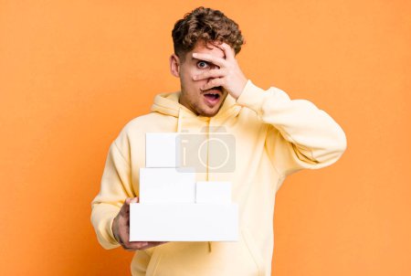Téléchargez les photos : Young adult caucasian man looking shocked, scared or terrified, covering face with hand. blank different packages concept - en image libre de droit