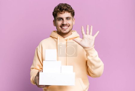 Foto de Young adult caucasian man smiling and looking friendly, showing number five. blank different packages concept - Imagen libre de derechos