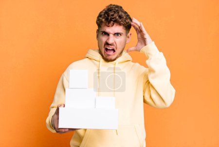 Foto de Young adult caucasian man screaming with hands up in the air. blank different packages concept - Imagen libre de derechos