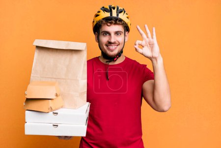 Foto de Young adult caucasian man feeling happy, showing approval with okay gesture.  take away fast food deliveryman concept - Imagen libre de derechos