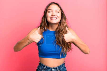 Photo for Young pretty woman smiling broadly looking happy, positive, confident and successful, with both thumbs up - Royalty Free Image