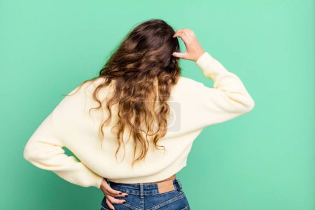 Photo for Young pretty woman feeling clueless and confused, thinking a solution, with hand on hip and other on head, rear view - Royalty Free Image